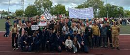 Grupowe zdjęcie drużyny OSP Stok wraz z kibicami. W przednim rzędzie kucają zawodnicy z medalami i pucharem, oraz dzieci, a za nimi stoją kibice uśmiechnięci i zadowoleni z osiągniętego miejsca w zawodach. Z prawej strony stoi Komendant Powiatowy PSP w Opocznie w piaskowym umundurowaniu koszarowym. Nad głowami zgromadzonych rozciągnięte są dwa transparenty z hasłami dopingującymi. W tle zielone boisko, drzewa oraz zachmurzone niebo. 