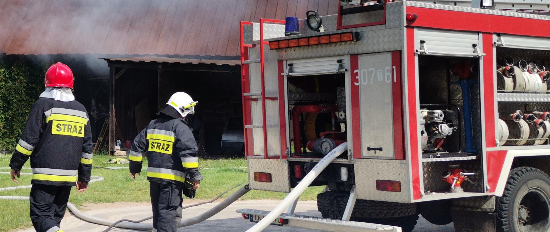 Zdjęcie przedstawia samochód gaśniczy biorący udział w akcji z podpiętymi wężami gaśniczymi. W tle budynek gospodarczy w którym zapalił się samochód.