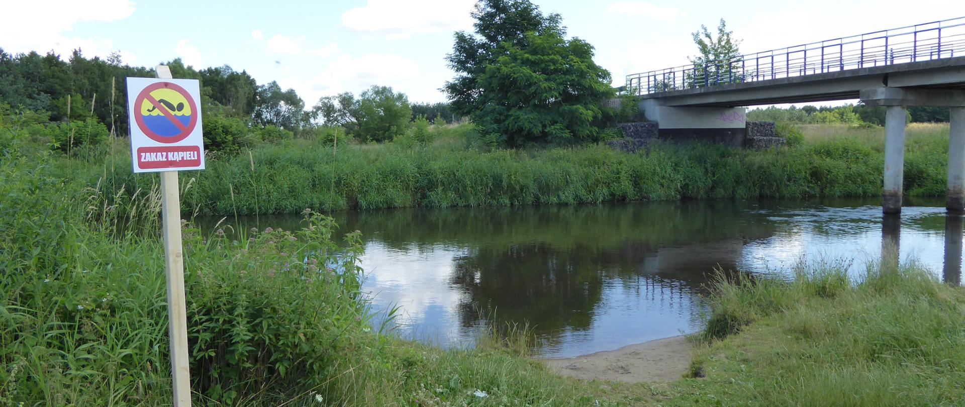 Zdjęcie przedstawia dzikie kąpielisko na rzece, na pierwszym planie widoczny znak zakaz kąpieli.