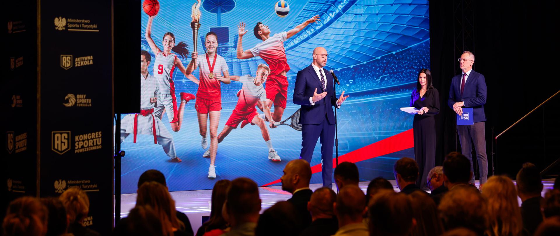 Zdjęcie przedstawiające ministra sportu i turystyki Jakuba Rutnickiego przemawiającego do mikrofonu na środku sceny podczas Kongresu Sportu Powszechnego 2025. Z prawej strony na scenie stoją prowadzący. Z lewej strony widać słup z logotypami MSiT, Orłów Sportu i Aktywnej Szkoły. Za plecami ministra grafika z dziećmi w strojach w biało-czerwonych barwach uprawiającymi różne sporty na stadionie PGE Narodowy. Na pierwszym planie widać widownię.