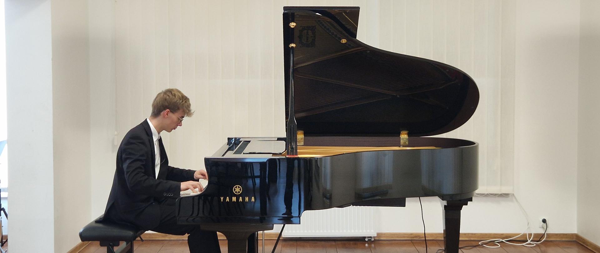 Na zdjęciu widać młodego mężczyznę w eleganckim stroju, grającego na czarnym fortepianie marki Yamaha. Mężczyzna siedzi z lewej strony kadru, zwrócony profilem w prawo, skupiony na klawiaturze. Ma krótkie, jasne włosy i okulary. Ubrany jest w ciemny garnitur z białą koszulą i krawatem w jasnym kolorze (prawdopodobnie biały lub srebrny).
Fortepian jest otwarty, co pozwala zobaczyć jego wnętrze i struny. Instrument zajmuje centralną i prawą część zdjęcia.
Tło stanowi jasna ściana z dużymi, białymi zasłonami lub żaluzjami, które rozciągają się przez całą szerokość ściany. Podłoga jest wykonana z ciemnego drewna o widocznych słojach. Po lewej stronie widać fragment jasnego korytarza lub innej części pomieszczenia. Oświetlenie jest jasne i równomierne, prawdopodobnie pochodzi z naturalnego światła wpadającego przez okna