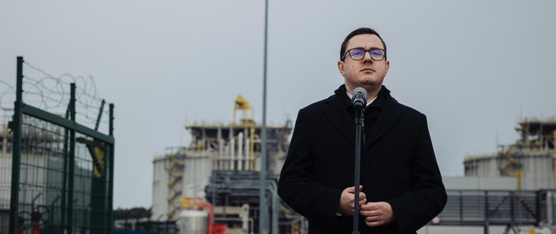 Minister Energii Miłosz Motyka przemawia stojąc przed mikrofonem. W tle zabudowania Terminala LNG w Świnoujściu