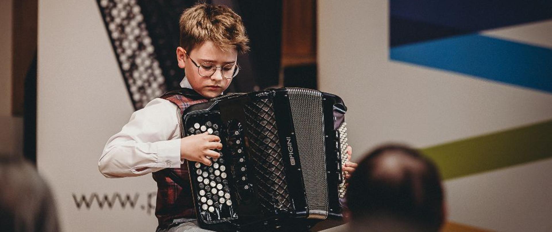 Zdjęcie przedstawia ucznia grającego na akordeonie. Na dole, w tle widoczny jest fragment publiczności.