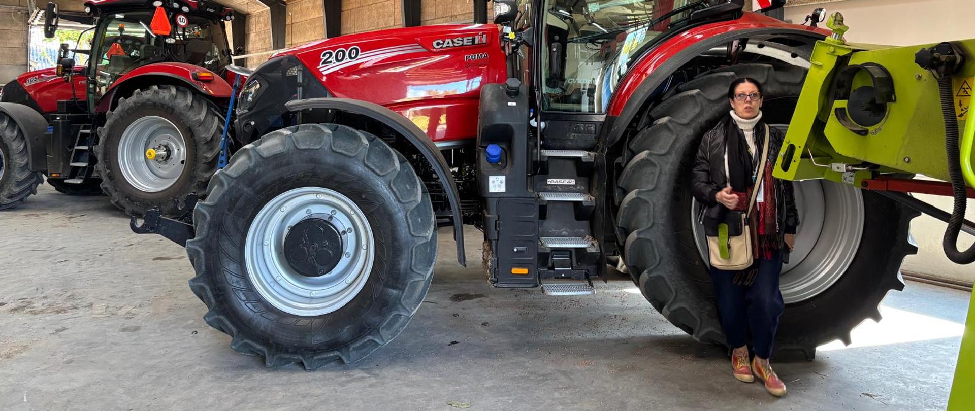 Uczestniczka seminarium EUROPEA stoi w hali maszyn rolniczych obok dużego czerwonego ciągnika Case IH. W tle widoczne są kolejne nowoczesne maszyny rolnicze.
