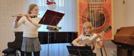 Uczennica gra na flecie, a obok Natalia Skrzydło gra na gitarze na Ogólnopolskim Kolędowym Konkursie Gitarowym w Bystrzycy Kłodzkiej - 12 stycznia 2024, w tle widoczny baner konkursowy