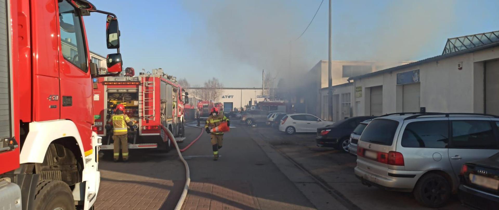 Po lewej stronie ustawione kolejno wozy strażackie, widziane od tyłu. Po prawej stronie zaparkowane pojazdy osobowe. Po środku, w tle, wydostające się kłęby dymu w pomieszczenia warsztatu samochodowego.