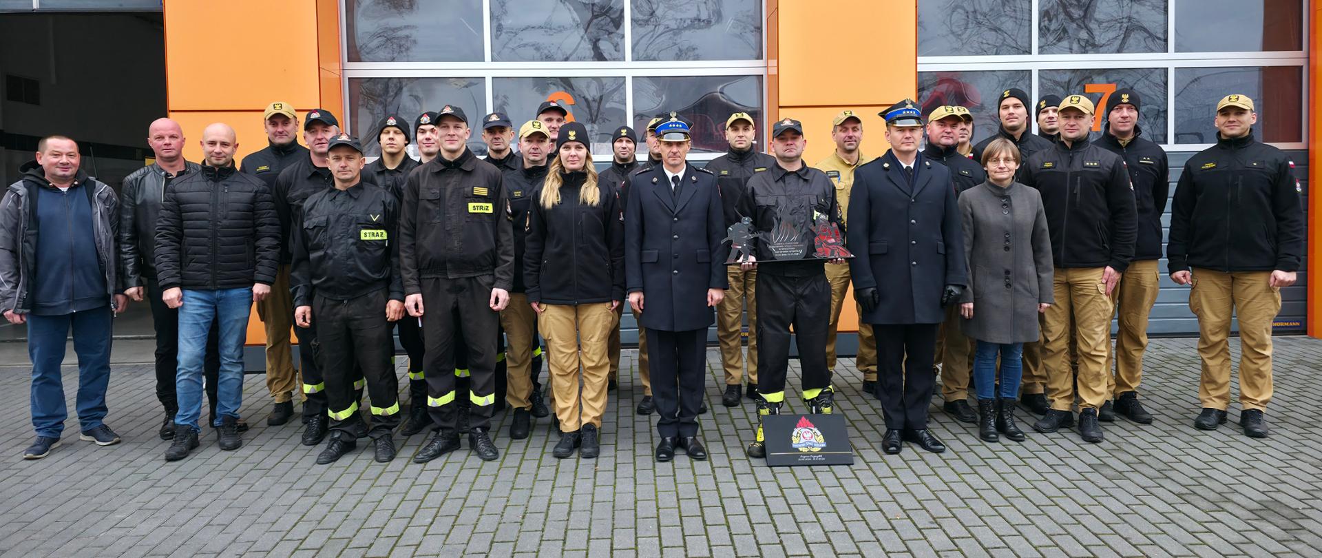 Na placu przed budynkiem strażnicy grupa strażaków w w umundurowaniu. Zdjęcie przedstawia moment uroczystej zbiórki. W otoczeniu stoją wozy i zabudowania jednostki.