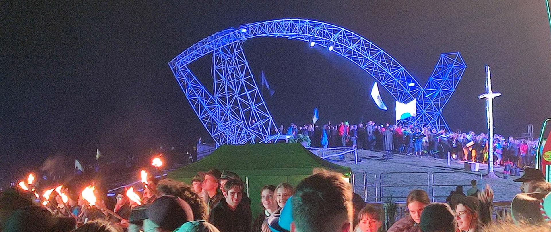 Noc. Na niebiesko podświetlona metalowa konstrukcja ryby. Wokół niej na wzgórzu przechodzący pod nią młodzi ludzie.