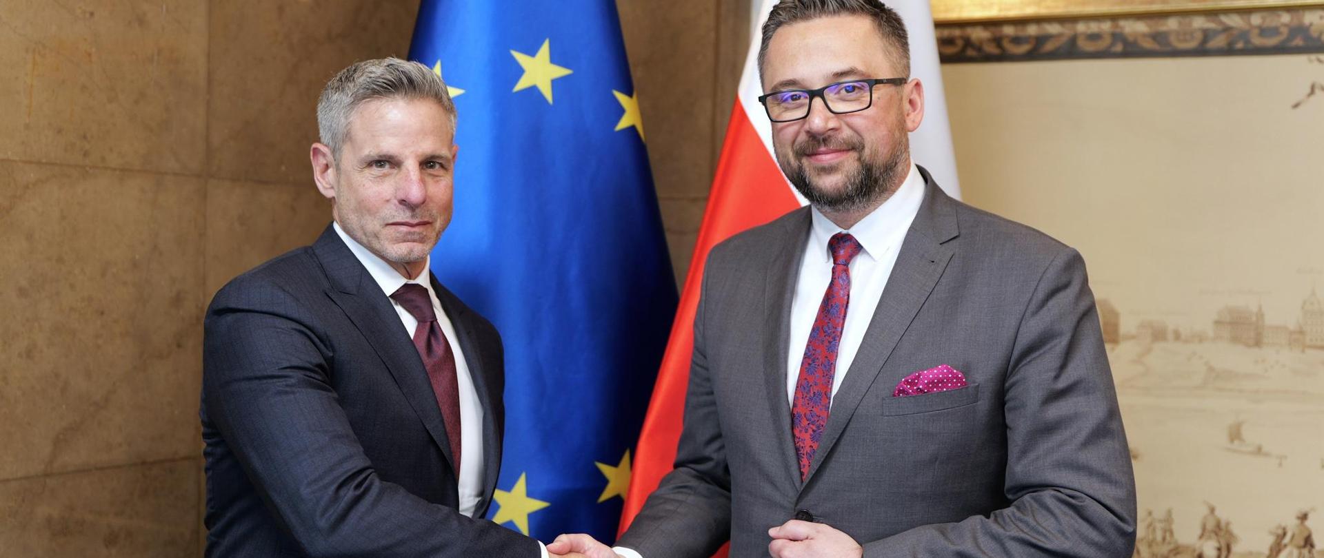 Dwóch mężczyzn podczas uścisku dłoni. Obaj ubrani w garnitury. W tle flaga UE i PL