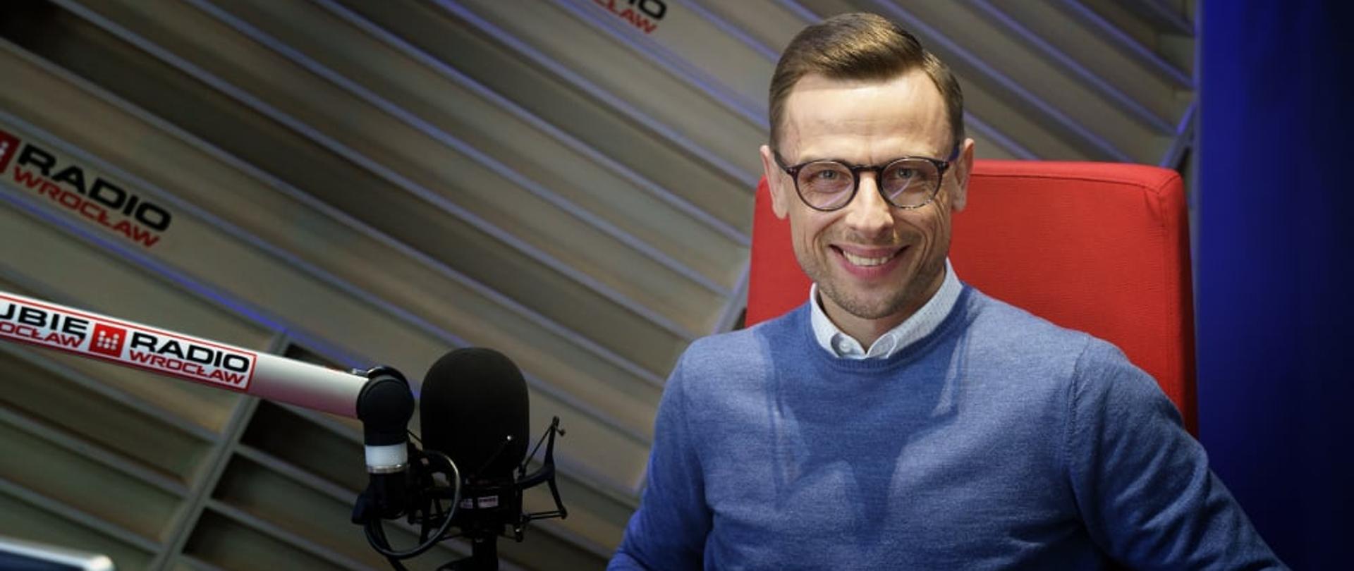 Dolnośląski Wojewódzki Inspektor Inspekcji Handlowej we Wrocławiu w studiu Radia Wrocław, mężczyzna ubrany jest w niebieski sweter, siedzi na czerwonym fotelu, przed nim na metalowym wysięgniku znajduje się mikrofon. 