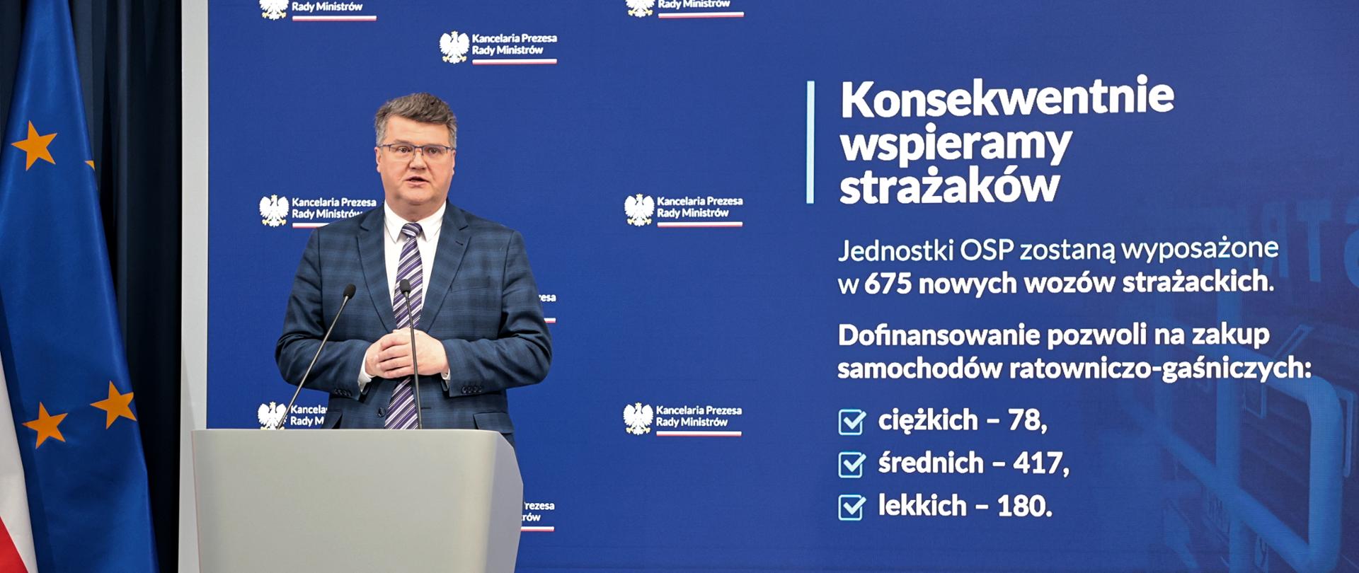 Osoba wypowiadająca się do mikrofonu, Za nią plansza z napisem Konsekwentnie wspieramy strażaków