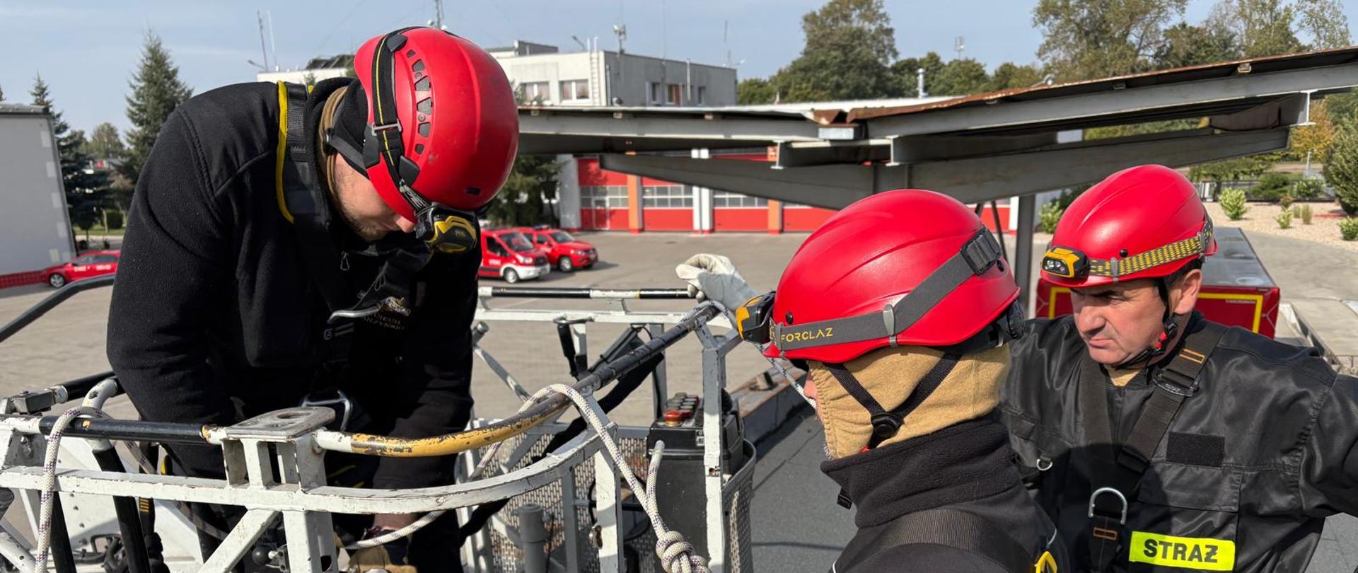 Strażacy w ekwipunku zabezpieczenia wysokościowego ćwiczą z kosza drabiny mechanicznej.