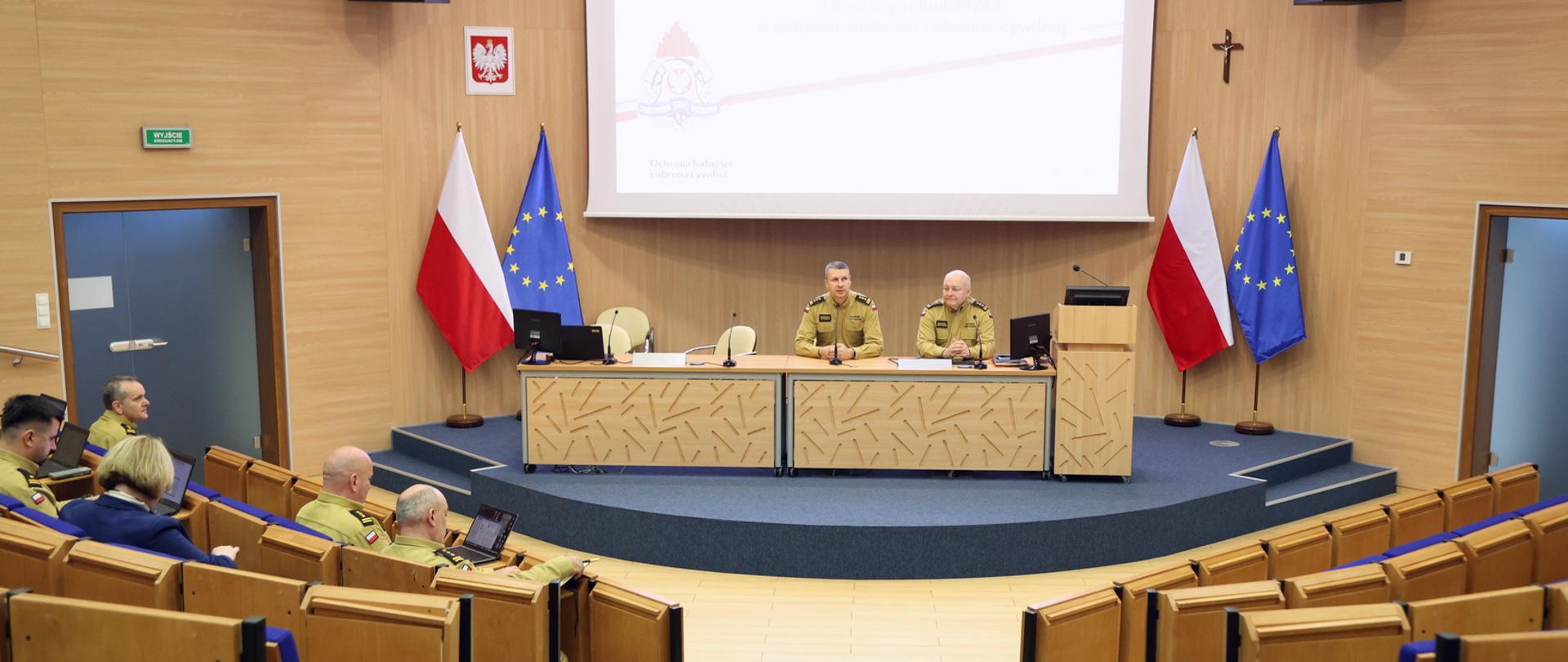 Widok z tyłu sali wykładowej podczas trwającej odprawy – na scenie dwóch funkcjonariuszy PSP prowadzi spotkanie zatytułowane „Odprawa służbowa i warsztaty”; obecni w sali korzystają z laptopów i notatek.