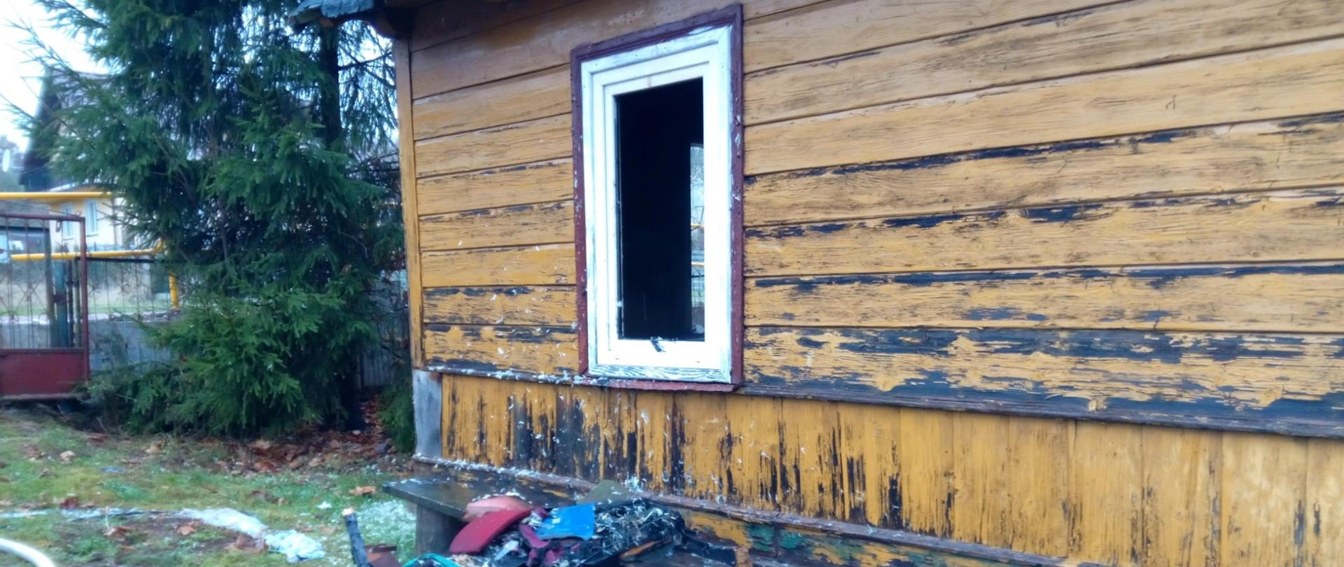 Zdjęcie przedstawia dom, gdzie doszło do pożaru. Widać wybite okno i nadpalone rzeczy usunięte z budynku.