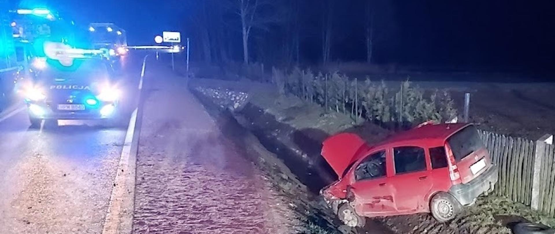 Samochód osobowy z uszkodzonym przodem stojący w przydrożnym rowie. Obok radiowóz z włączonymi sygnałami świetlnymi.