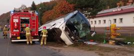 Zdjęcie zrobione na zewnątrz, podczas usuwania skutków wypadku. Na boku leży autokar, obok niego stoi samochód gaśniczy, strażacy prowadzą akcję ratowniczą. Przednia szyba autokaru jest rozbita.