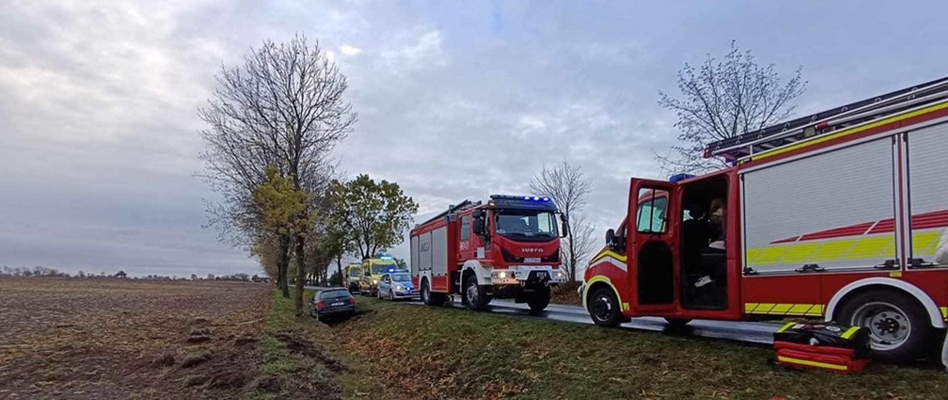 Na zdjęciu znajdują się 2 wozy strażackie, samochód policyjny oraz 2 zespoły ratownictwa medycznego. W rowie znajduje się samochód osobowy.