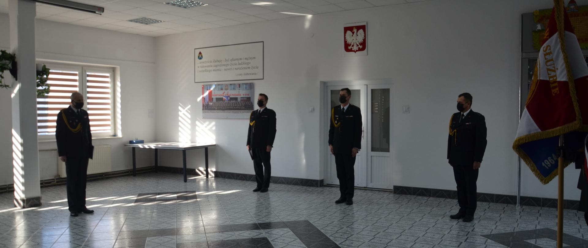W dużym pomieszczeniu stoi czterech strażaków w umundurowaniu galowym. Strażacy stoją w znacznych odległościach od siebie. Po prawej stronie widoczny jest sztandar na drzewcu. 