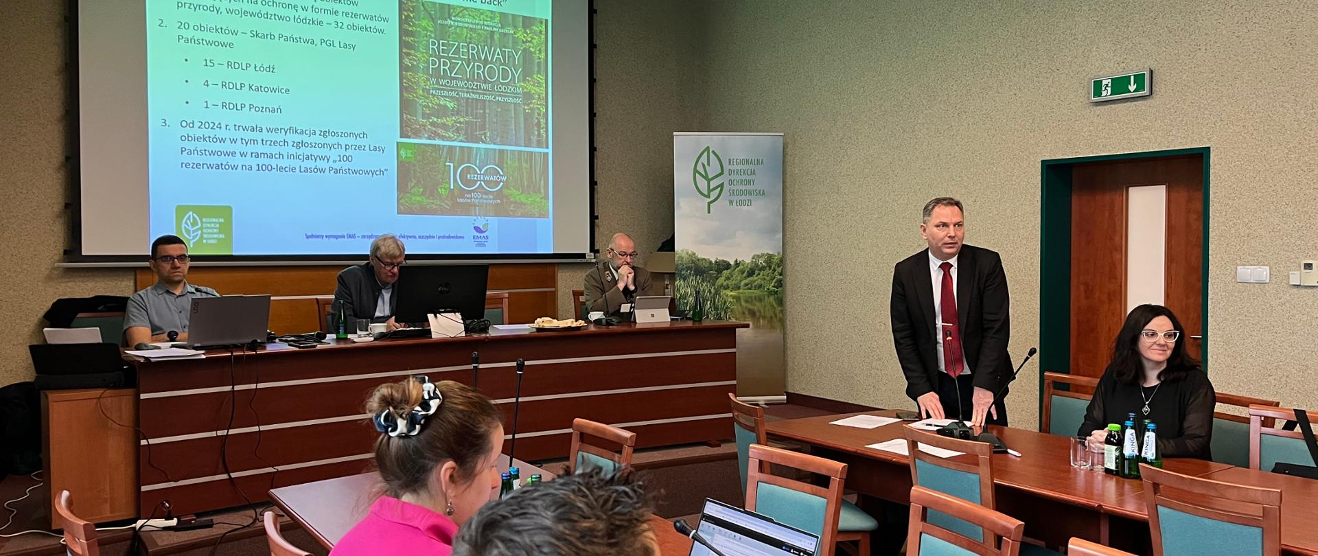 Sala konferencyjna podczas posiedzenia – przy stole prezydialnym siedzą uczestnicy z laptopami, a mężczyzna w garniturze przemawia przy mównicy. W tle widoczny slajd prezentacji dotyczący rezerwatów przyrody, na sali obecni są uczestnicy spotkania