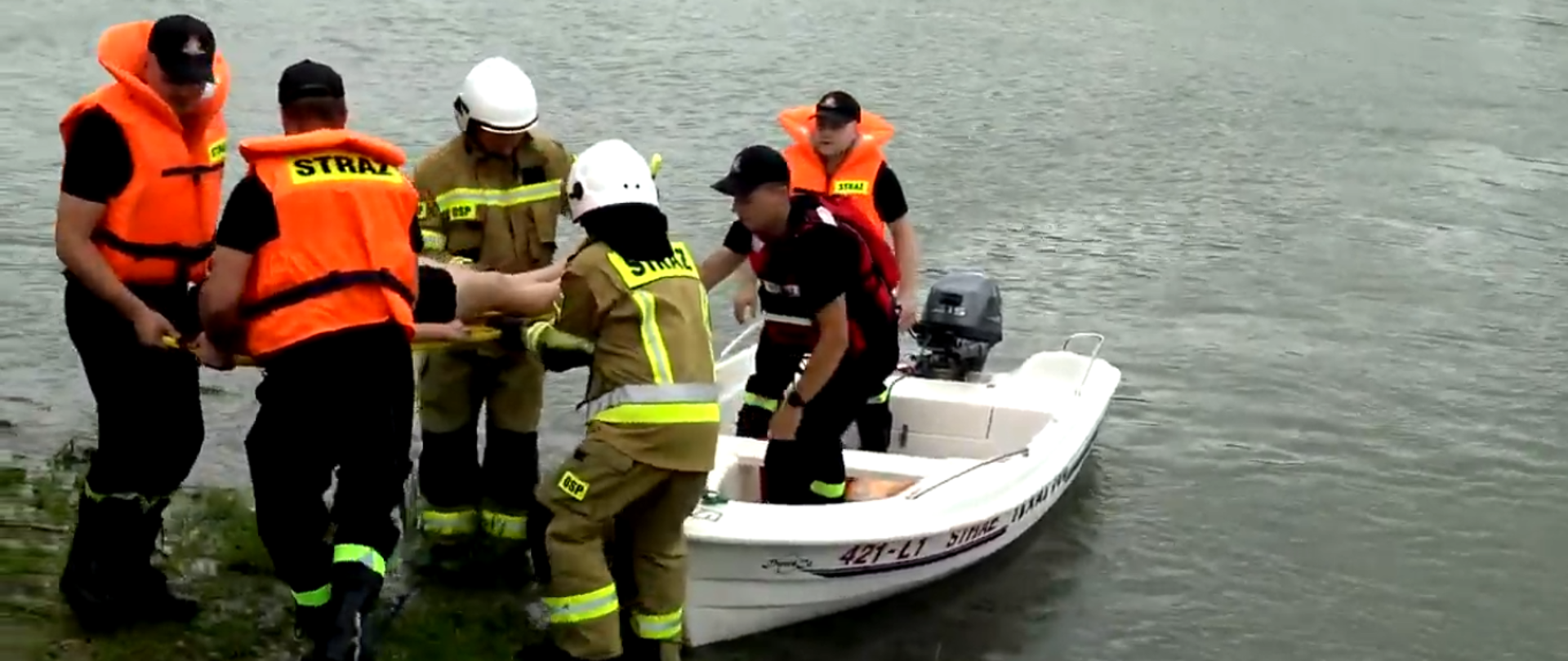 Zbiornik wodny a na nim łódka w kolorze białym z napisem straż. W łódce dwaj strażacy w kamizelkach ratunkowych. Na brzegu przed łódką czterej strażacy trzymający deskę ratowniczą . Na desce znajduje się osoba poszkodowana.