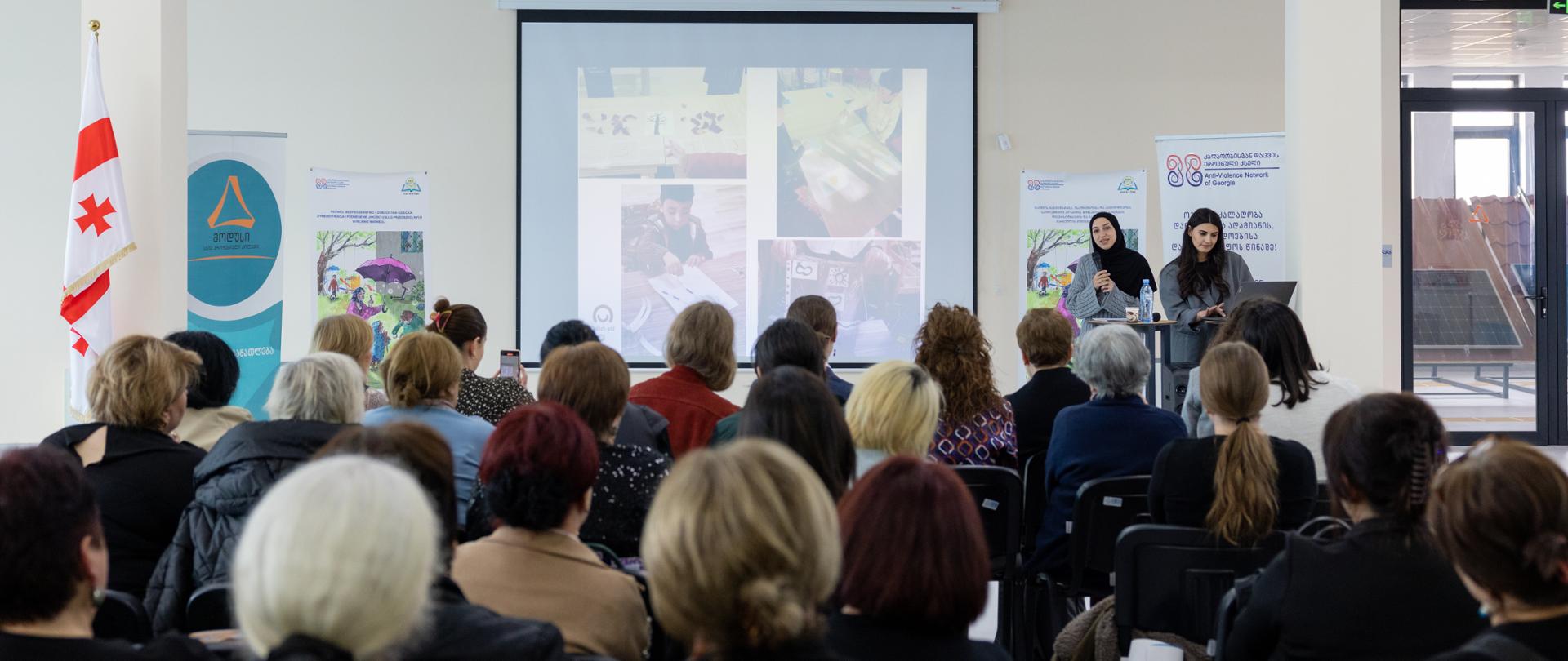 Sala konferencyjna z grupą osób, prezentują dwie kobiety, na prezentacji widać zdjęcia z warsztatów dla dzieci