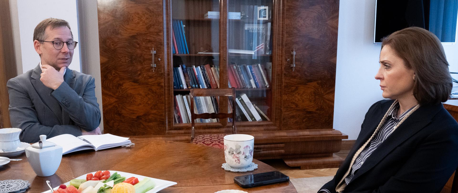 Minister Joanna Mucha siedzi w gabinecie przy stole z Bartłomiejem Balcerzykiem, rozmawiają. 