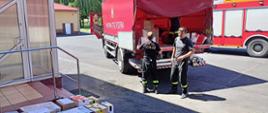 Na zdjęciu strażacy ładują na samochód ciężarowy z czerwoną plandeką paczki w kartonach.
