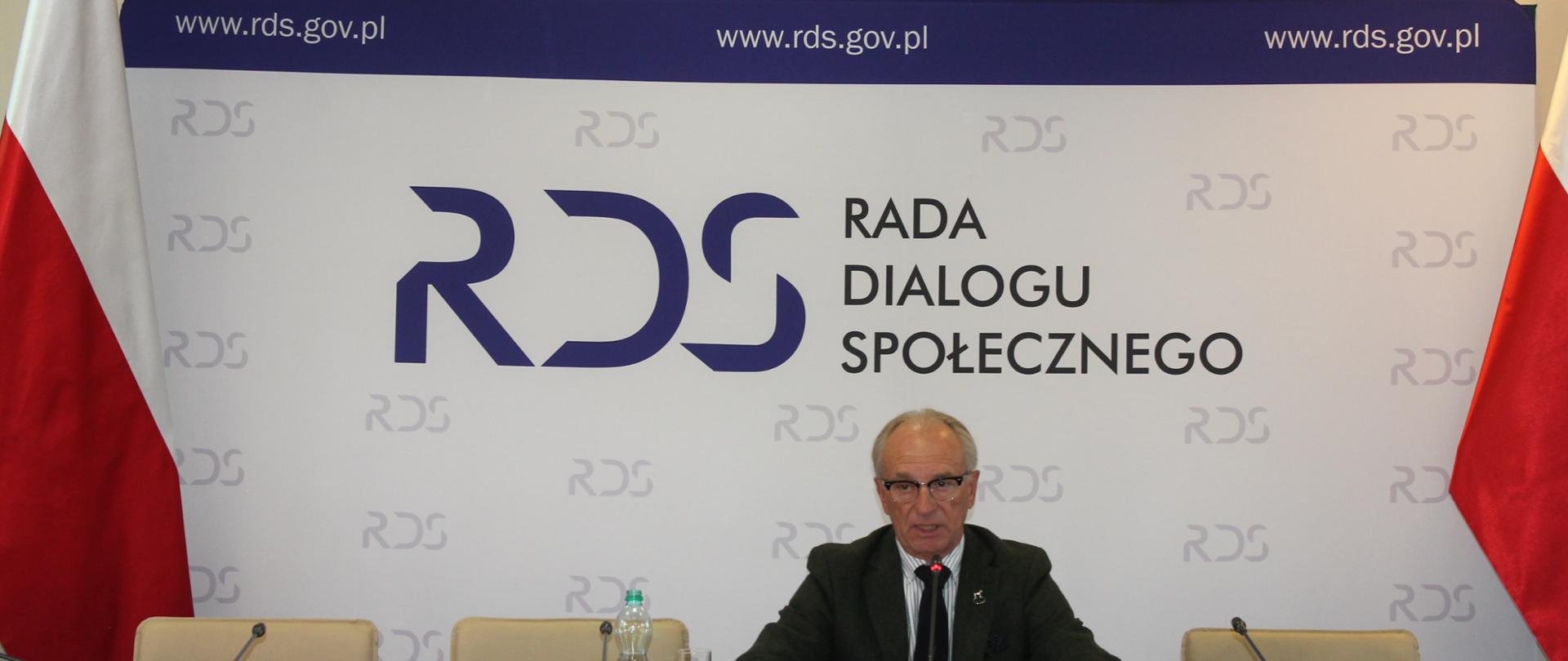 Zdjęcie przedstawia stół prezydialny za którym siedzi Pan Marek Kowalski – Przewodniczący Podzespołu ds. zamówień publicznych. Po obu stronach stołu znajdują się flagi Polski. W tle znajduje się baner z napisem Rada Dialogu Społecznego. Nad banerem znajduje się Godło Polski.