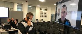 Funkcjonariusz Państwowej Straży Pożarnej rozmawia przez telefon komórkowy, na przeciwko telewizor w którym wyświetla się osoba prowadząca ćwiczenia. Niedaleko funkcjonariusze siedzą przy komputerach.