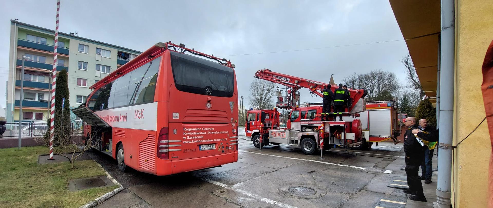 Autobus Centrum Krwiodawstwa i Krwiolecznictwa w Szczecinie zaparkowany na parkingu przed Komendą Straży pożarnej w Gryfinie. W tle wóz strażacki z wysięgnikiem. 
