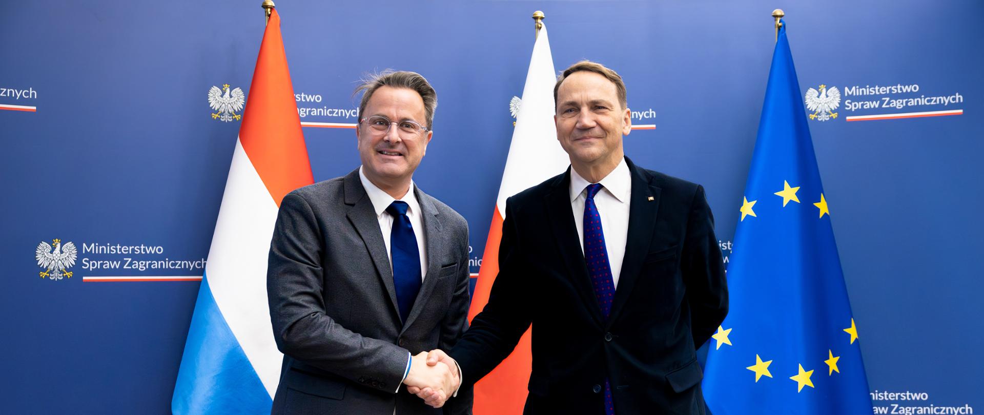 Polish and Luxembourg foreign ministers standing side by side during an official meeting, with national flags and an official backdrop behind them. 