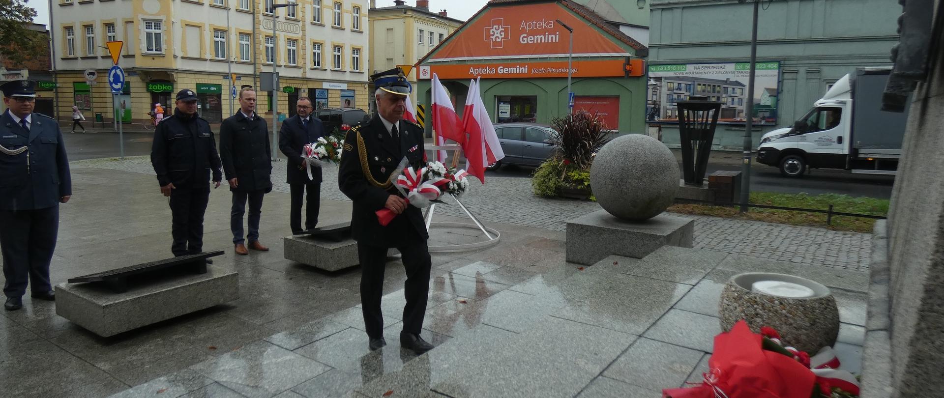 Uczczenie rocznicy. Plac przed pomnikiem Żołnierza Polskiego w Rawiczu. Delegacja rawickich służb mundurowych składa wiązankę kwiatów. Funkcjonariusz Państwowej Straży Pożarnej trzymając w rękach wiązankę kwiatów wchodzi po schodkach w pobliże pomnika, gdzie leżą już inne, wcześniej złożone kwiaty. Pozostali członkowie delegacji stoją kilka metrów za strażakiem. Z prawej strony stojak z flagami państwowymi. W tle budynki, przechodnie i ruch uliczny.