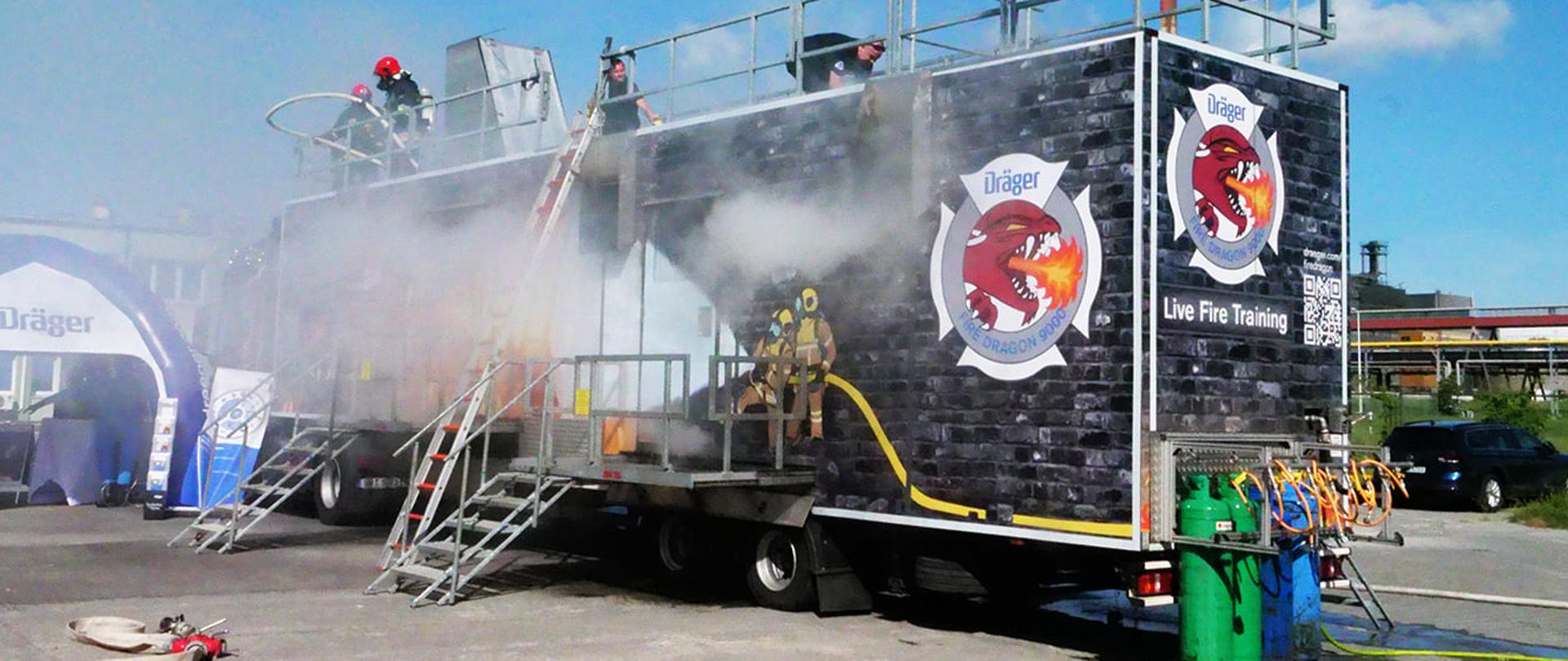Na zdjęciu widoczny czarny kontener z logo ziejącej ogniem głowy smoka, namalowanymi strażakami gaszącymi pożar, z napisem Fire Dragon 9000 i białym napisem Live Fire Training. Na dachu zabezpieczonym stalową barierką widać dwóch mężczyzn w czarnych koszulkach, za nimi w głębi dwóch strażaków w czarnych ubraniach bojowych i czerwonych hełmach na głowie, u jednego z nich na plecach widoczna butla. Nad dachem kontenera widoczny komin z wydobywającym się białym dymem, przed kontenerem metalowe podesty ze schodami i metalowa drabina oparta o dach. Przed kontenerem widoczne stalowe zielone i niebieskie zbiorniki stojące na wilgotnym betonowym placu. Z lewej strony zdjęcia rozwinięty jasny, popielaty wąż gaśniczy, dalej biało-niebieski pneumatyczny namiot. Z prawej strony widoczny tył i prawy bok samochodu typu kombi i tereny zielone, dalej elementy infrastruktury zakładu z zabudowaniami.