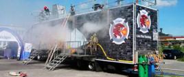 Na zdjęciu widoczny czarny kontener z logo ziejącej ogniem głowy smoka, namalowanymi strażakami gaszącymi pożar, z napisem Fire Dragon 9000 i białym napisem Live Fire Training. Na dachu zabezpieczonym stalową barierką widać dwóch mężczyzn w czarnych koszulkach, za nimi w głębi dwóch strażaków w czarnych ubraniach bojowych i czerwonych hełmach na głowie, u jednego z nich na plecach widoczna butla. Nad dachem kontenera widoczny komin z wydobywającym się białym dymem, przed kontenerem metalowe podesty ze schodami i metalowa drabina oparta o dach. Przed kontenerem widoczne stalowe zielone i niebieskie zbiorniki stojące na wilgotnym betonowym placu. Z lewej strony zdjęcia rozwinięty jasny, popielaty wąż gaśniczy, dalej biało-niebieski pneumatyczny namiot. Z prawej strony widoczny tył i prawy bok samochodu typu kombi i tereny zielone, dalej elementy infrastruktury zakładu z zabudowaniami.