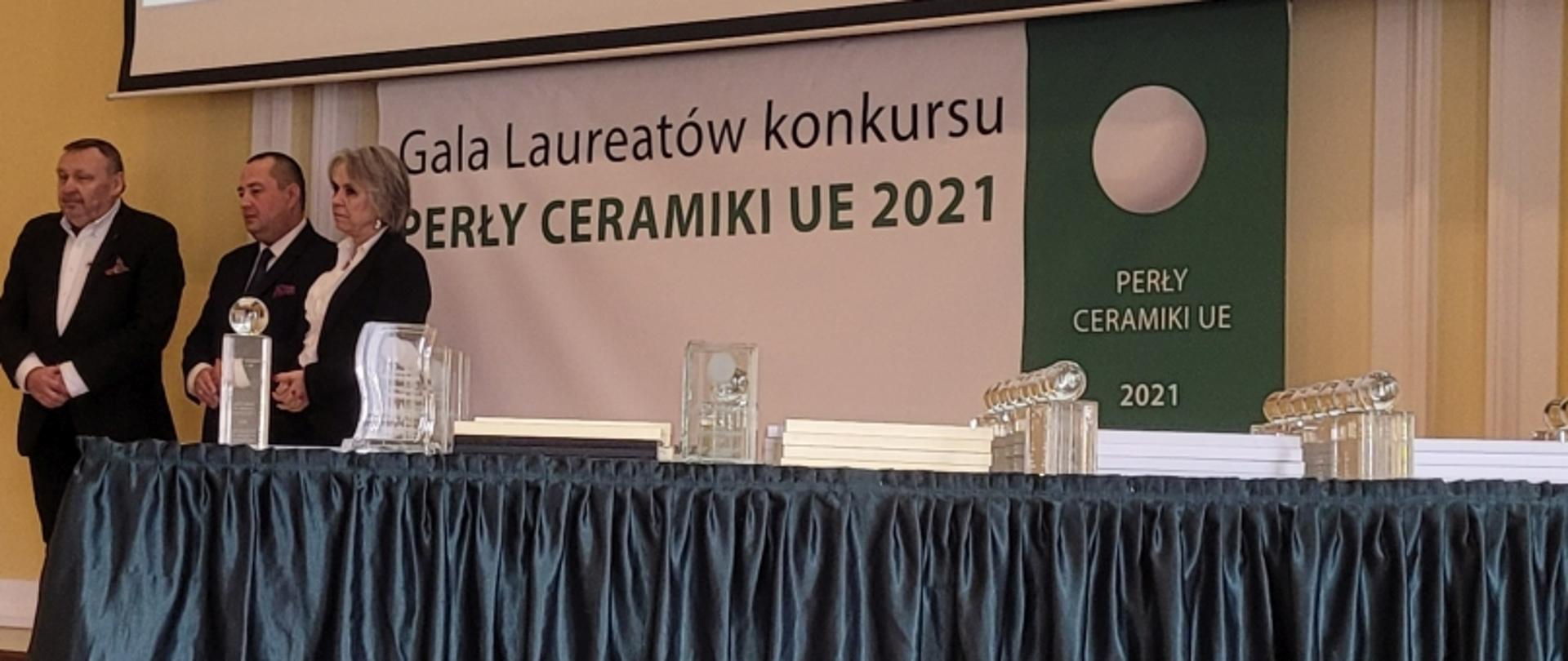 Uroczystość wręczenia nagród „PERŁY CERAMIKI UE 2021” — trzy osoby na scenie, baner w tle, trofea na stole z granatowym obrusem.