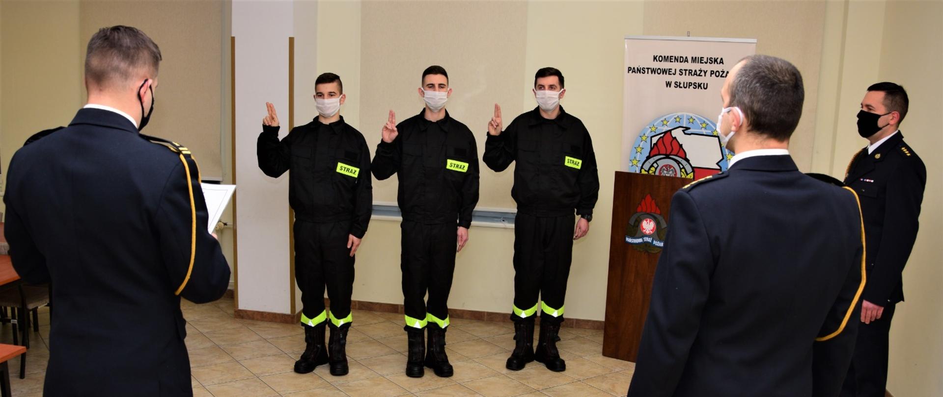 Zdjęcie przedstawia ślubowanie strażaków w sali w KM PSP w Słupsku. Ślubowanie odbywa się w obecności Komendanta Miejskiego oraz dowódców JRG. Na zajęciu trzech strażaków w ubranych koszarowych, Komendant oraz dowódcy w mundurach galowych.
