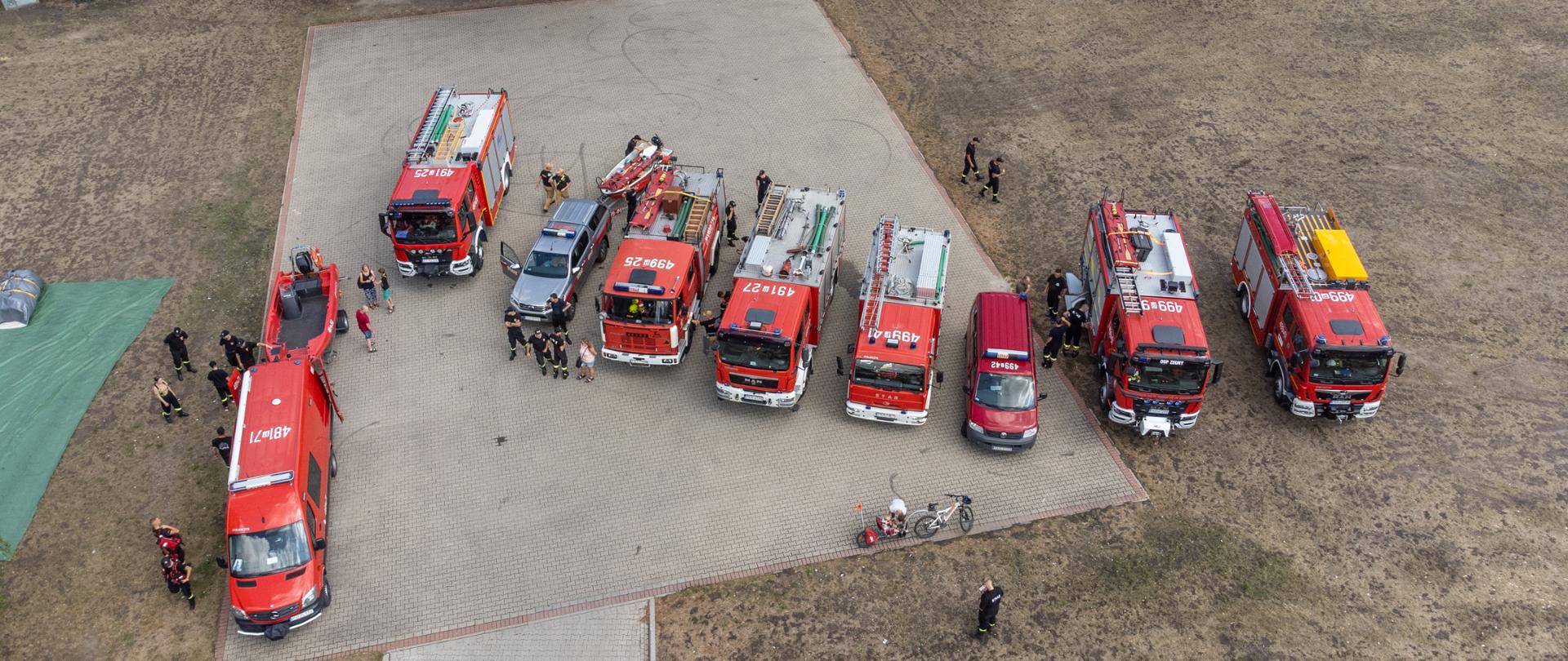 Na placu manewrowym stoją pojazdy pożarnicze usytuowane w szeregu. Rozpoczynają się warsztaty z ratownictwa wodnego dla jednostek KSRG. Trwa omówienie przebiegu warsztatów. 
