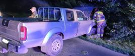 Pick-up marki Nissan częściowo wjechany w zarośla przy drodze, nocą. Strażak w ubraniu ochronnym i odblaskach pochylony nad otwartą maską pojazdu. Obok tylnej części auta stoi mężczyzna w czapce z daszkiem, oświetlony niebieskim światłem służb ratunkowych.