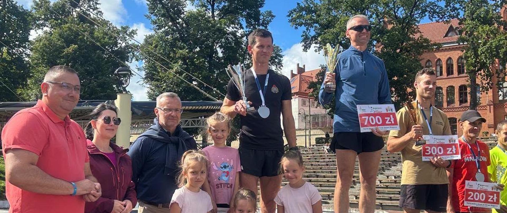 Na zdjęciu widać ceremonię wręczenia nagród podczas zawodów sportowych. Trzech mężczyzn stoi na podium, trzymając puchary i nagrody pieniężne, a obok nich pozują organizatorzy oraz dzieci. Wydarzenie odbywa się na świeżym powietrzu, przy słonecznej pogodzie, na tle drzew i zabytkowych budynków.