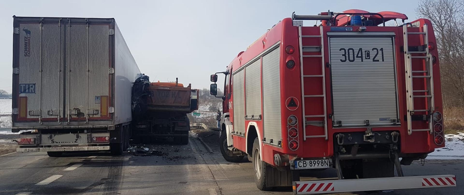 Od lewej strony widać dwa tyły aut ciężarowych, pierwsze z lewej naczepa koloru białego, drugie naczepa koloru pomarańczowego. Od prawej strony widać tył auta ciężarowego strażackiego koloru czerwonego z srebrnymi roletami. 