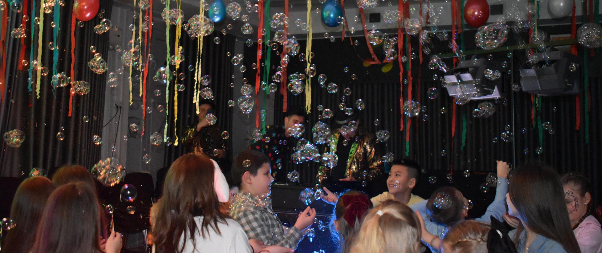 Na zdjęciu znajdują się tańczące dzieci, u góry zawieszone są kolorowe balony, bibuła, bańki.