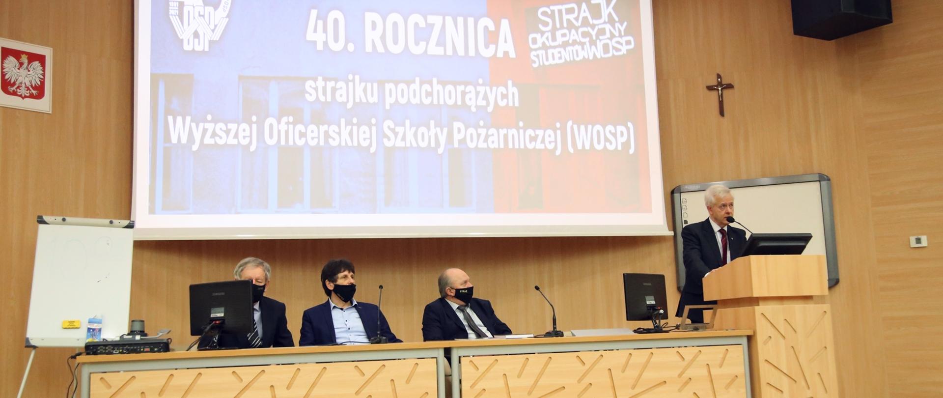 Wnętrze auli, na podeście przy mównicy przemawia jeden z honorowych gości, pozostałych trzech gości siedzi przy biurku. W tle rozwinięty ekran na którym widoczna jest prezentacja