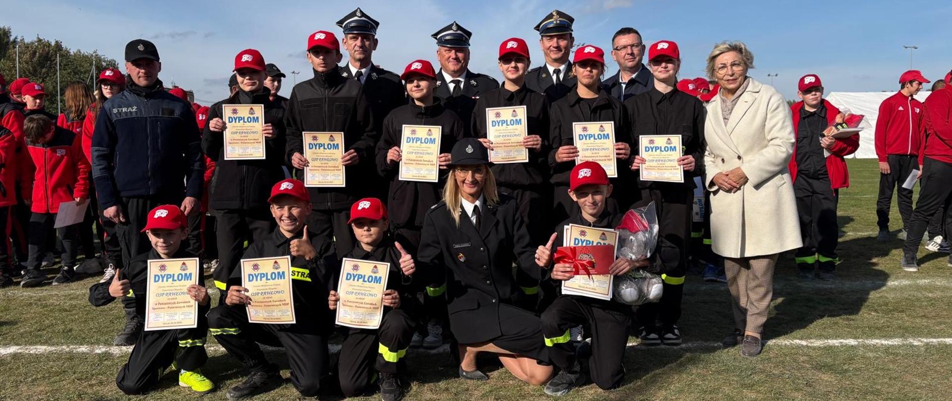 Zdjęcie przedstawia grupę młodych strażaków i ich opiekunów podczas uroczystości na świeżym powietrzu. Wszyscy pozują do wspólnego zdjęcia, trzymając dyplomy i nagrody. Uczestnicy ubrani są w mundury strażackie i czerwone czapki z logo OSP (Ochotnicza Straż Pożarna). W centrum grupy, w pierwszym rzędzie, znajduje się kobieta w mundurze, klęcząca z przodu razem z młodymi strażakami. Po prawej stronie stoi elegancko ubrana kobieta w jasnym płaszczu,. W tle widać więcej uczestników w czerwonych kurtkach oraz namioty, co sugeruje, że wydarzenie ma miejsce podczas zawodów lub uroczystości strażackiej na otwartym terenie. Pogoda jest słoneczna, a niebo niemal bezchmurne.