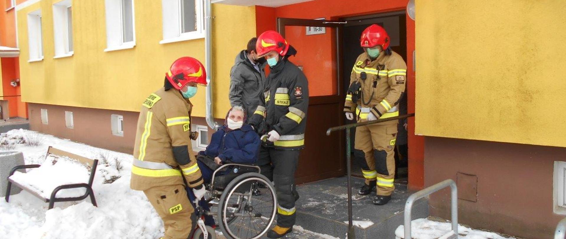 Zdjęcie przedstawia strażaków PSP wynoszących starszą kobietę na wózku inwalidzkim z klatki schodowej bloku