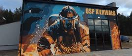 Budynek remizy OSP Kierwałd z dużym muralem przedstawiającym strażaka w akcji, z napisem „OSP KIERWAŁD” nad wejściem.