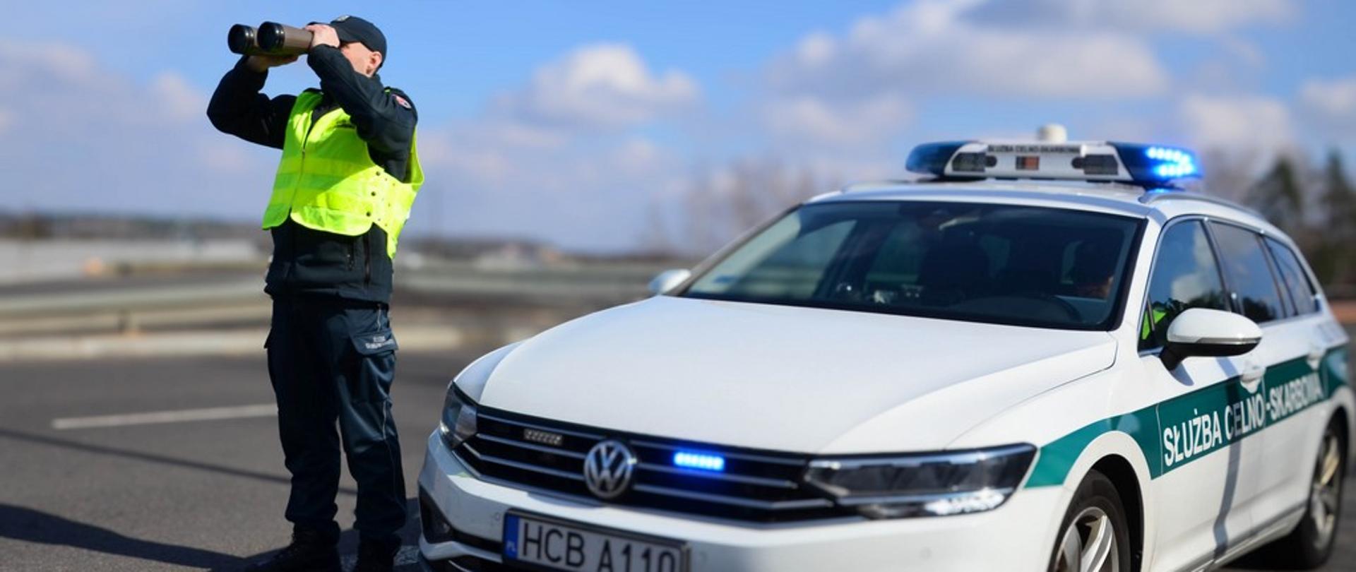 Funkcjonariusz patrzy przez lornetkę stojąc obok radiowozu