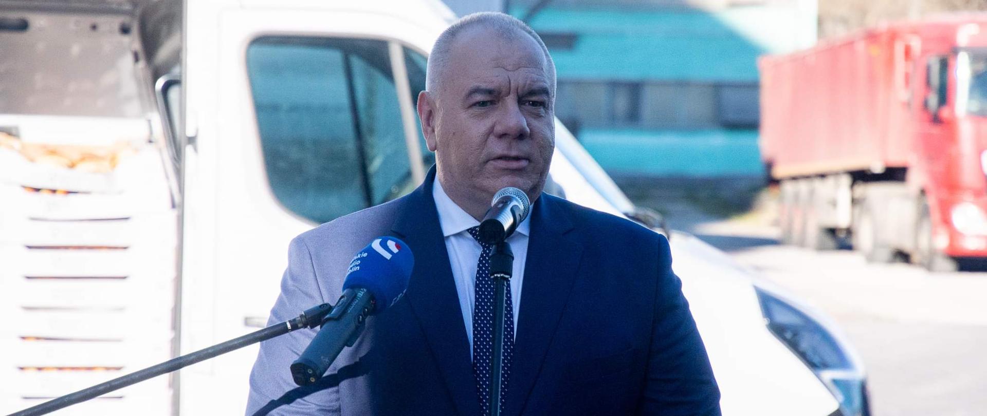 Wicepremier Jacek Sasin na tle samochodu z darami Zamojski Zakładów Zbożowych dla żołnierzy. W tle samochód dostawczy zakładów zbożowych w Zamościu. Przed premierem stoją mikrofony