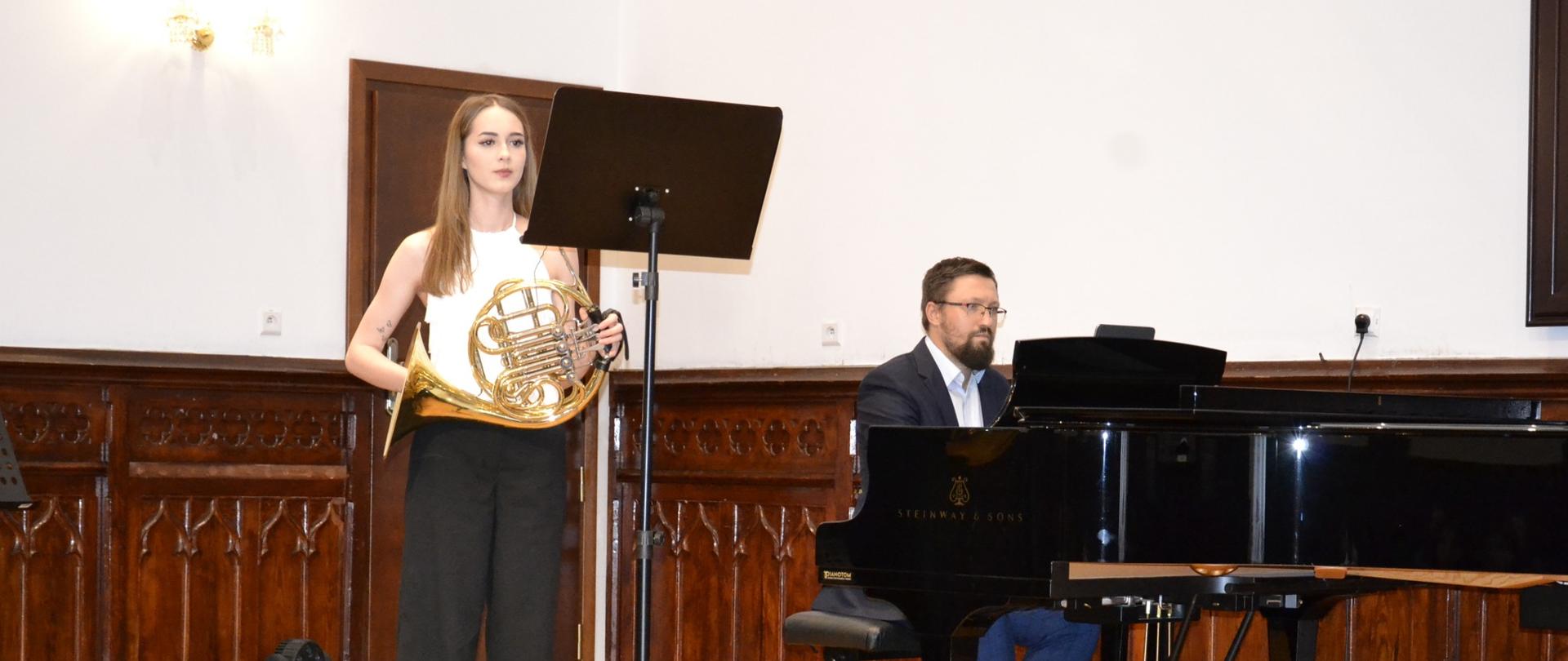 Uczennica trzymająca waltornię stoi przy pulpicie nutowym, a nauczyciel akompaniuje na fortepianie Steinway podczas recitalu dyplomowego w Sali Królewskiej.