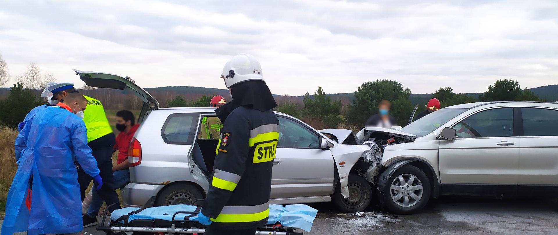 Zdjęcie przedstawia dwa samochody osobowe które zderzyły się czołowo. Z prawej strony widać ratownika medycznego i policjanta - na pierwszym planie widać strażaka. Przed samochodami leżą nosze.