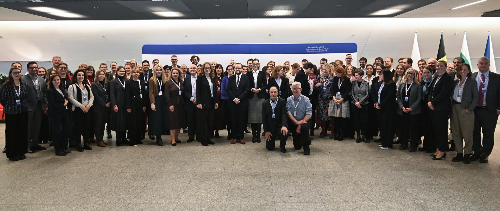 Zdjęcie zbiorowe, pod białą ścianą na szarej podłodze stoi duża grupa kilkudziesięciu osób.